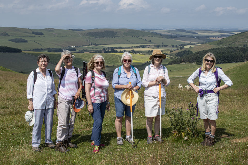 A group of people stand in a line on a grassy hillside with a view to distant fields and hills